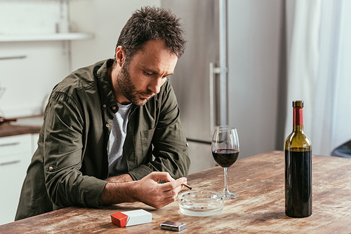 Disappointed man smoking cigarette beside wine bottle and glass on kitchen table