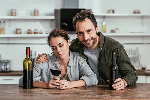 Smiling man with beer hugging sad woman with wine on kitchen