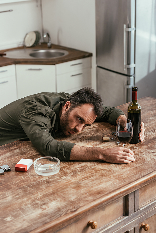 Drunk man holding wine bottle and glass beside cigarettes on kitchen table