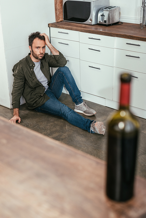 Selective focus of man with alcohol addiction sitting on floor and looking at wine bottle on kitchen table