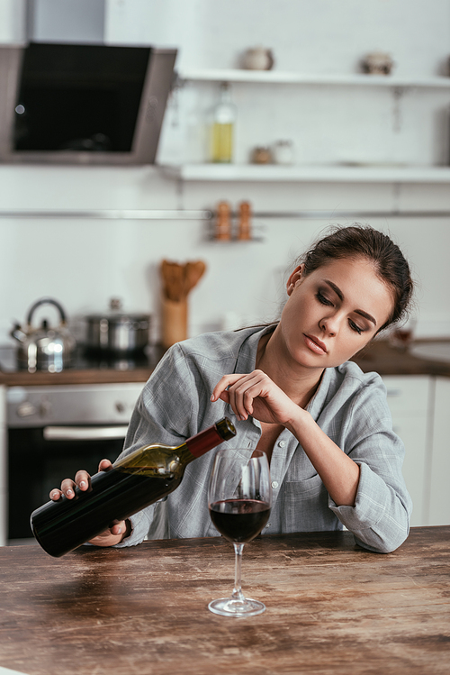 Sad woman with alcohol addicted pouring wine in glass on kitchen