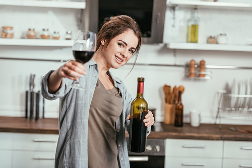 Selective focus of smiling woman holding bottle and wine glass at kitchen