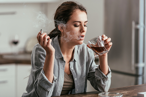 Woman smoking and looking at whiskey glass at kitchen table