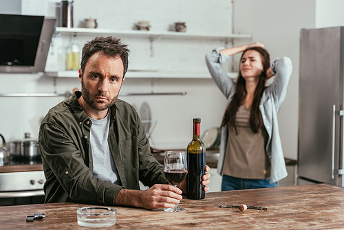 Selective focus of alcohol addicted man with wine and upset wife on kitchen