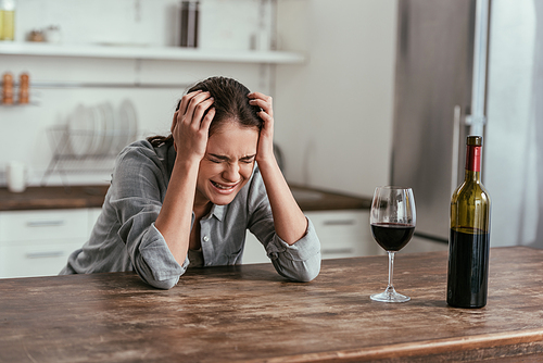 Woman crying beside wine glass and bottle on kitchen table