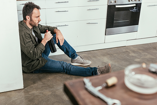 Selective focus of sad man with wine bottle sitting on kitchen floor