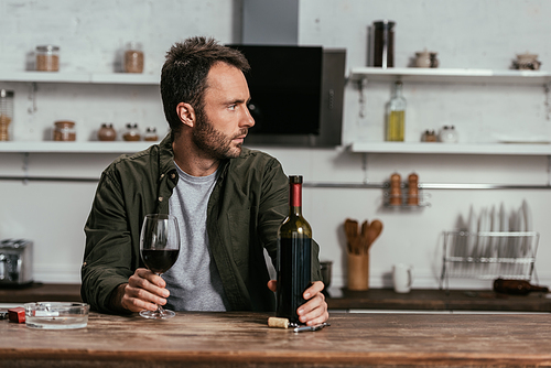 Man with wine glass and bottle looking away at kitchen table