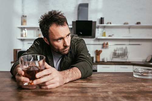 Selective focus of worried man holding whiskey glass beside ashtray on kitchen table