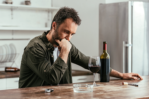 Pensive man looking at wine glass and bottle on kitchen table