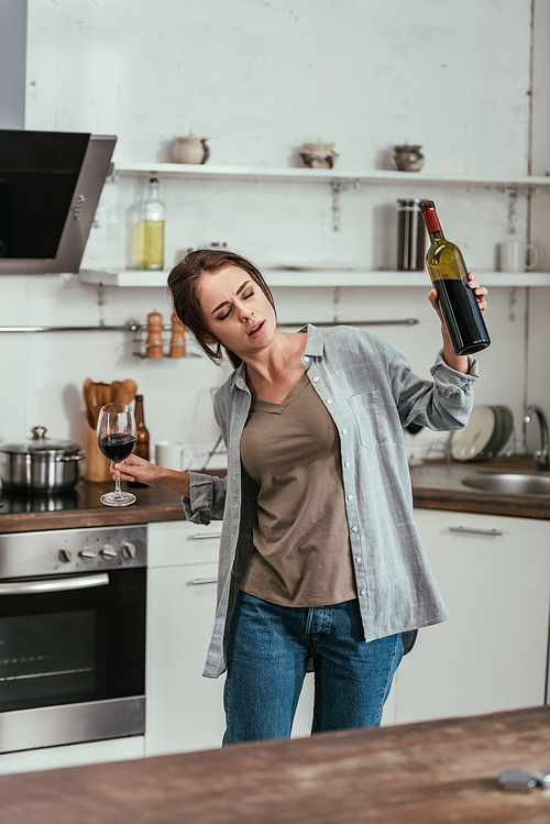 Woman with alcohol depended holding wine glass and bottle on kitchen