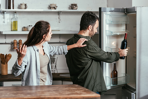 Angry woman quarreling with husband taking wine bottle from fridge