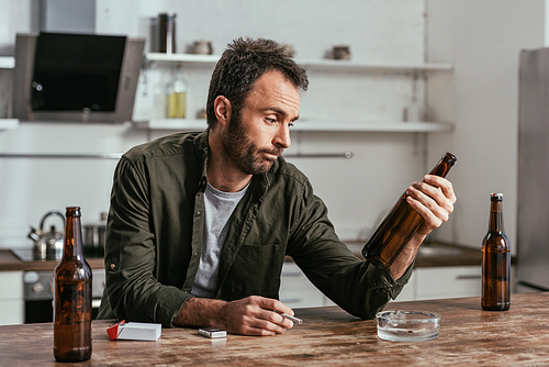 Sad man with alcohol depended holding cigarette and beer bottle at table
