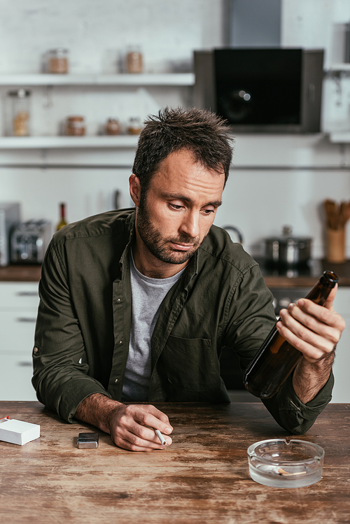 Upset man with alcohol depended holding cigarette and beer bottle at table