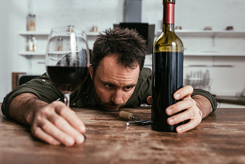 Selective focus of sad man holding wine glass and bottle on kitchen table