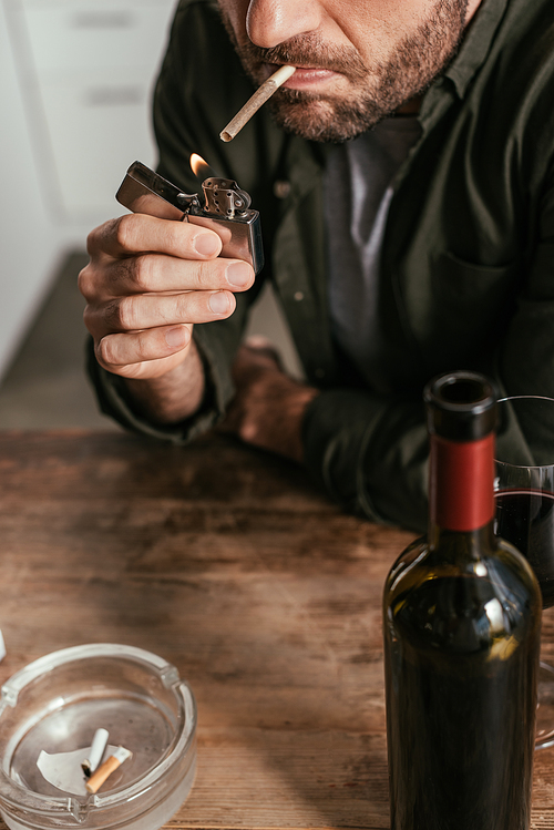 Cropped view of man lighting cigarette beside wine glass and bottle on table