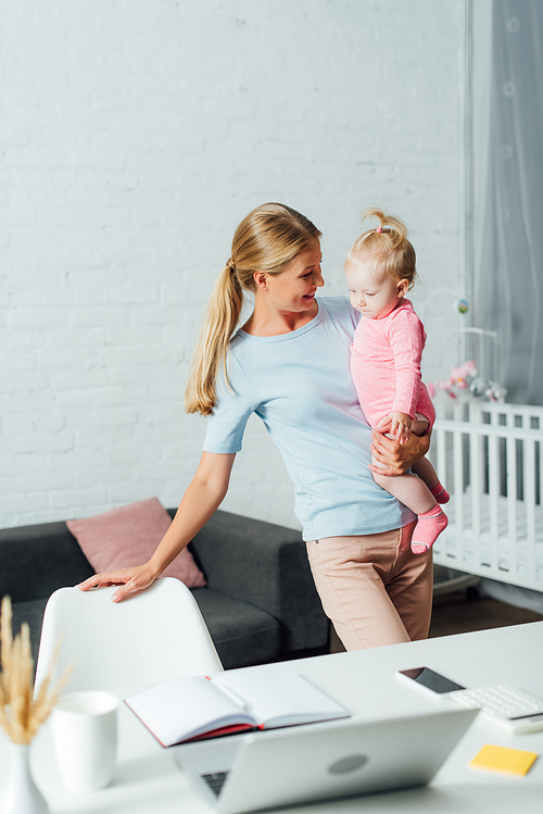 Selective focus of mother holding baby daughter near gadgets and notebook on table at home