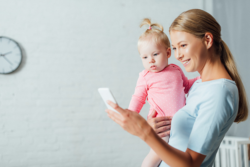 Selective focus of woman using smartphone while holding baby daughter at home