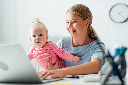 Selective focus of mother holding baby girl and using laptop at home