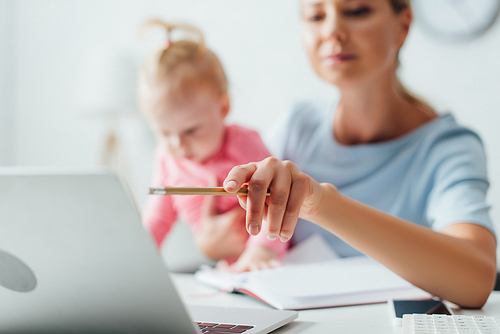 Selective focus of woman pointing with pencil at laptop while holding infant girl at home