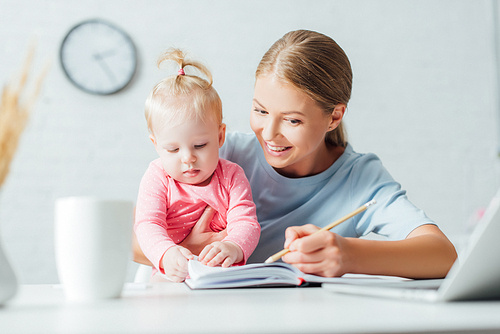 Selective focus of woman writing on notebook and holding baby daughter while working at table