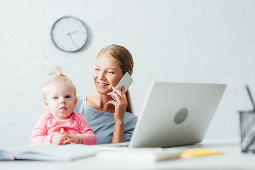 Selective focus of woman talking on smartphone while working near baby daughter at home