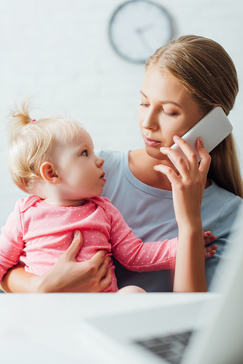 Selective focus of woman talking on smartphone and looking at infant daughter near laptop on table