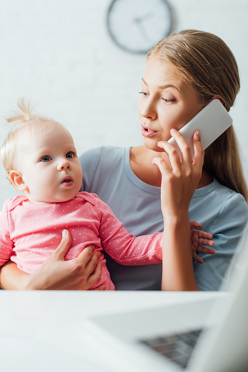 Selective focus of mother holding baby girl and talking on smartphone while working at home