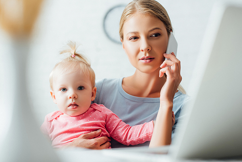 Selective focus of focused woman talking on smartphone and holding daughter at home