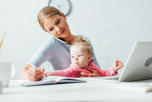 Selective focus of infant girl sitting near mother talking on smartphone and writing on notebook at home