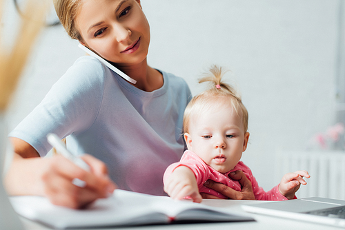 Selective focus of woman talking on cellphone while working near baby daughter at table