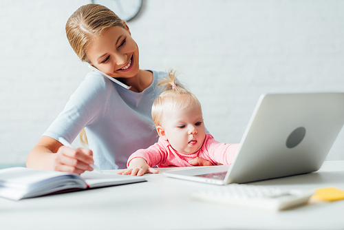 Selective focus of infant sitting near mother talking on smartphone and working at home