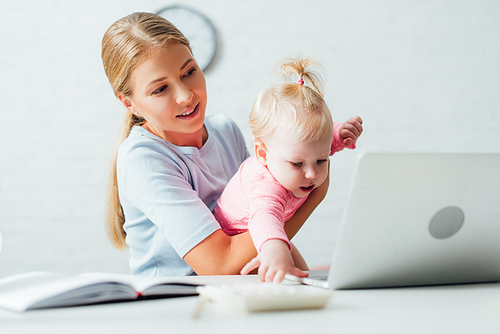 Selective focus of woman holding infant daughter while working with notebook and laptop at home