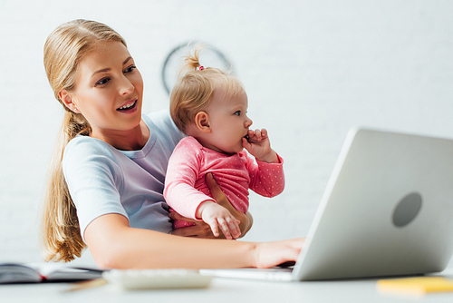 Selective focus of mother holding baby girl while working on laptop at home