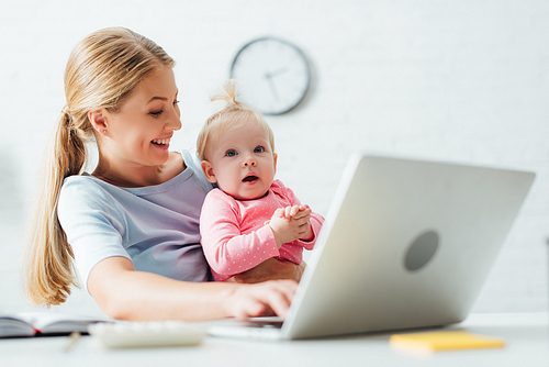 Selective focus of mother looking at baby girl while working with laptop at table