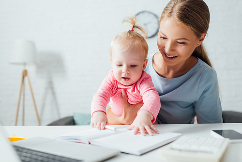 Selective focus of infant touching notebook near mother and gadgets on table