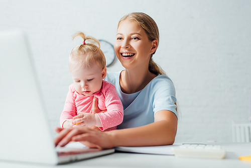 Selective focus of woman holding daughter and using laptop at table