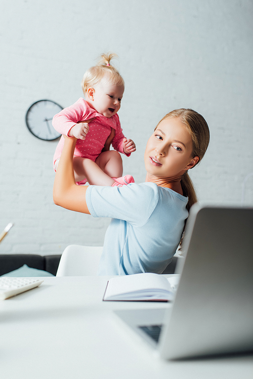 Selective focus of woman playing with baby girl while looking at laptop on table at home