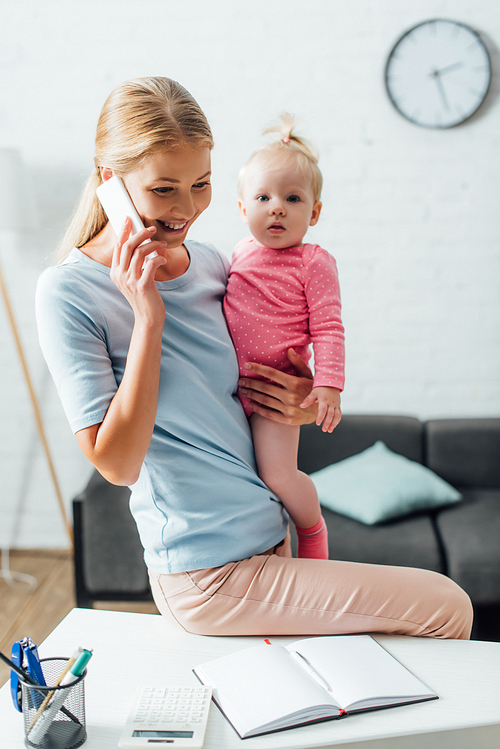 Selective focus of woman talking on smartphone and holding baby girl near stationery on table