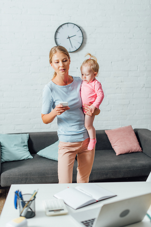 Selective focus of woman holding smartphone and baby girl near stationery and laptop on table