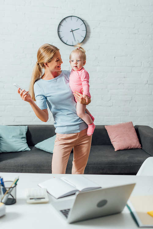 Selective focus of woman holding smartphone and looking at child at home