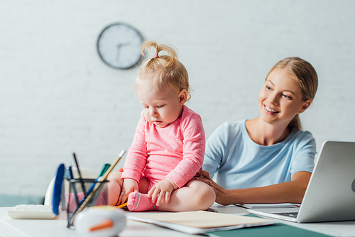 Selective focus of baby girl sitting near stationery and laptop on table beside mother