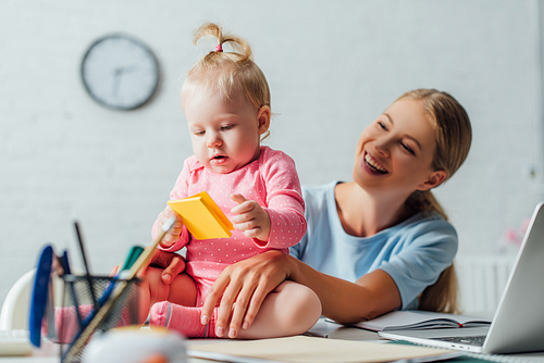 Selective focus of mother looking at baby girl holding sticky notes near laptop on table