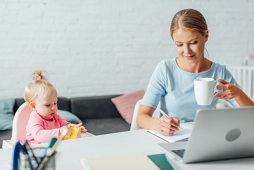 Selective focus of infant girl holding sticky notes near mother with cup writing on notebook at home