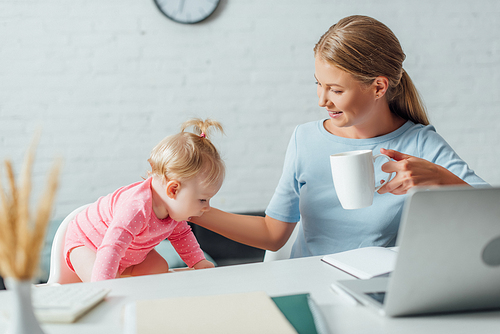 Selective focus of woman holding cup and touching baby girl near laptop and stationery on table