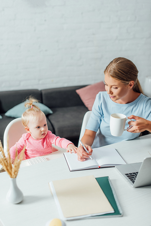 Selective focus of baby girl sitting near mother holding cup and writing on notebook at home