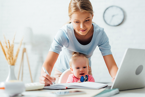 Selective focus of woman writing on notebook while working near infant with stapler at table