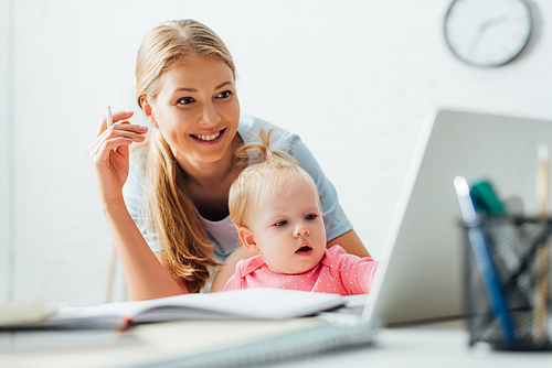 Selective focus of woman holding pen and looking at laptop near child at home