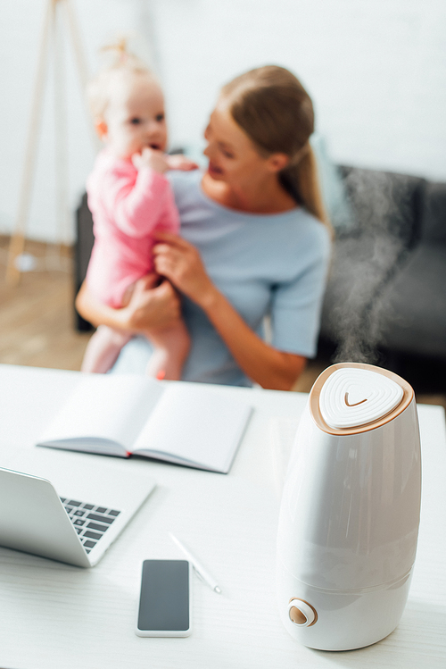 Selective focus of humidifier near gadgets on table and mother holding infant girl at background