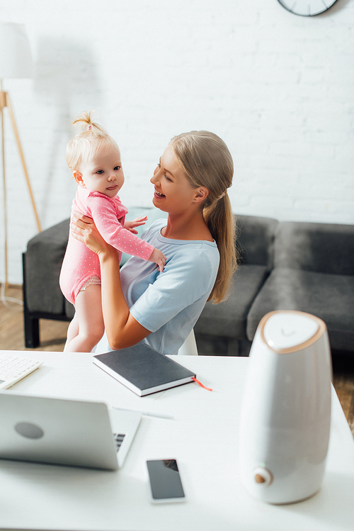 Selective focus of mother holding baby girl near gadgets, notebook and humidifier on table