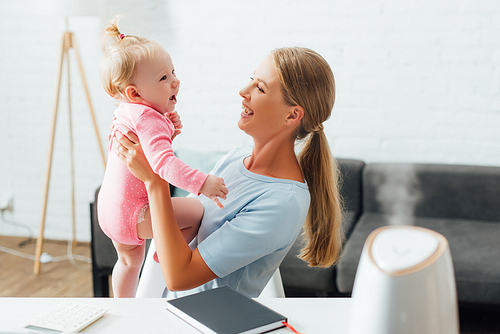 Selective focus of woman holding infant near notebook and humidifier on table